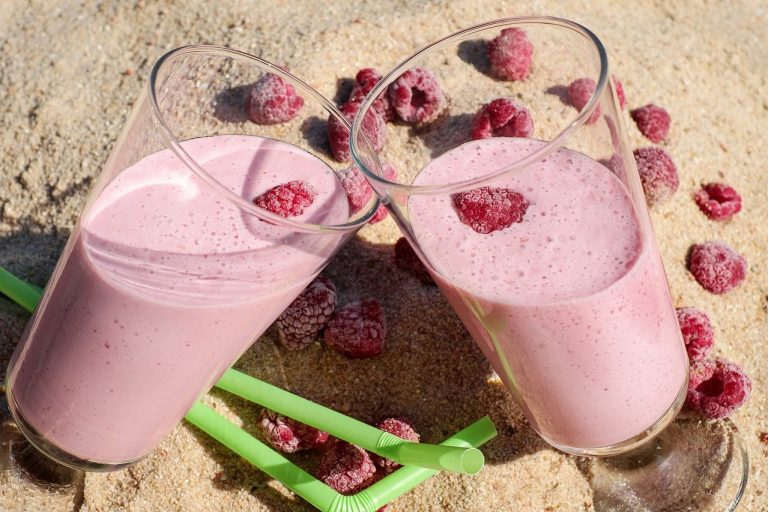 Zwei Gläser Himbeersmoothie mit frischen Himbeeren auf sandigem Untergrund.